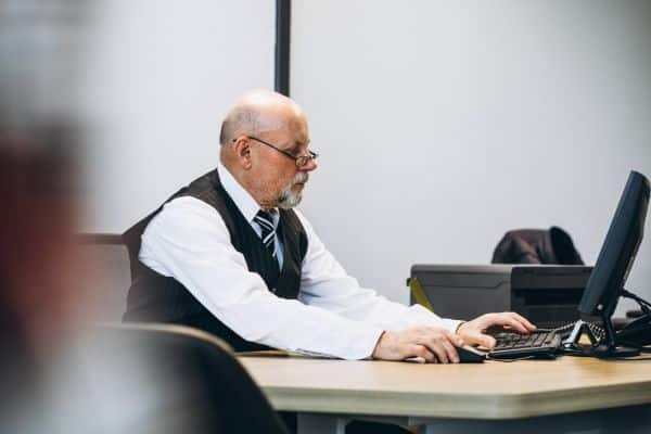 An older man in a suit and vest sits at a desk, focused on using a computer.